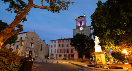 Historic square Place Mirabeau in French town of Pertuis overlooking illuminated statue with fountain on background with medieval donjon with clock and parish church of Saint-Nicolas on summer evening - obrazy, fototapety, plakaty