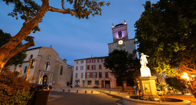 Historic square Place Mirabeau in French town of Pertuis overlooking illuminated statue with fountain on background with medieval donjon with clock and parish church of Saint-Nicolas on summer evening