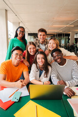 Vertical portrait. Group of young teenage students siting at library, using a laptop and looking at camera. Happy classmates studying and doing homeworks at high school. Multiracial academy people