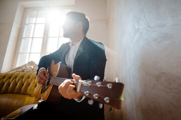 Low angle view of handsome young man playing guitar in the modern hotel room with sunlight