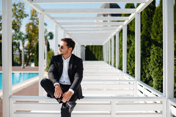 Handsome young man in suit and sunglasses sitting on white stairs near the pool at sunny day and looking away