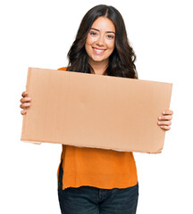 Beautiful brunette young woman holding cardboard blank empty banner looking positive and happy standing and smiling with a confident smile showing teeth