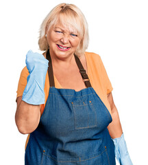 Middle age blonde woman wearing cleaner apron and gloves angry and mad raising fist frustrated and...
