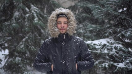 A young man stands against a background of fir trees under heavy snowfall in winter. A man in a...