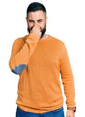 Hispanic man with beard wearing casual winter sweater smelling something stinky and disgusting, intolerable smell, holding breath with fingers on nose. bad smell
