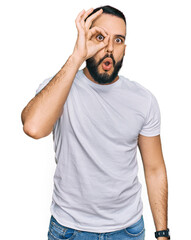 Fototapeta premium Young man with beard wearing casual white t shirt doing ok gesture shocked with surprised face, eye looking through fingers. unbelieving expression.
