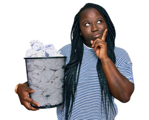 Young black woman with braids holding paper bin full of crumpled papers serious face thinking about question with hand on chin, thoughtful about confusing idea