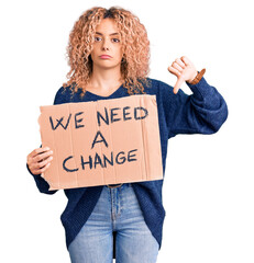 Young blonde woman with curly hair holding we need a change banner with angry face, negative sign...