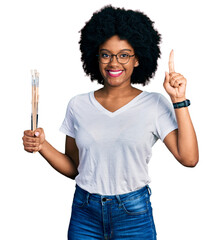 Young african american woman holding paintbrushes smiling with an idea or question pointing finger...