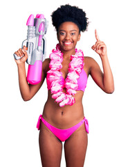 Young african american woman wearing bikini and hawaiian lei holding water gun surprised with an...