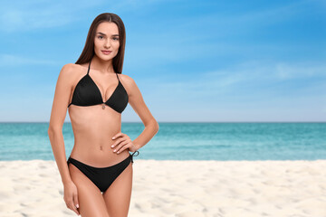 Beautiful woman in stylish black bikini on sandy beach near sea, space for text