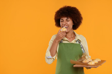 Happy young woman in apron holding board with cookies on orange background. Space for text