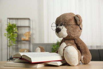 Teddy bear with glasses and open books on wooden table in room