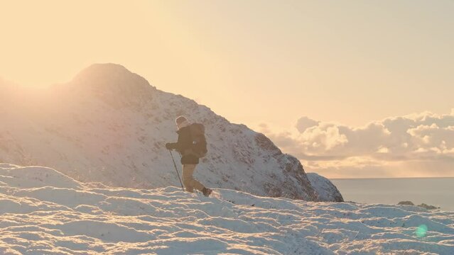 The mountaineering spirit comes alive as a hiker, adorned with a backpack, conquers the snowy ascent towards the mountain's pinnacle. A breathtaking winter sunset bathes the landscape in golden hues.