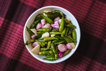 pickle of whole chili pepper, shallot and garlic in a white bowl with red bamboo braid n the background