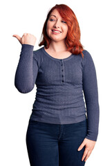 Obraz premium Young beautiful redhead woman wearing casual t-shirt smiling with happy face looking and pointing to the side with thumb up.
