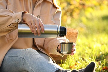 Woman pouring tea from thermos into cup lid on green grass outdoors, closeup