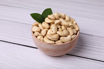 Tasty cashew nuts and green leaves in bowl on light wooden table, closeup