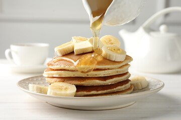 Fototapeta na wymiar Pouring honey from jug onto delicious pancakes with bananas and butter at white wooden table, closeup