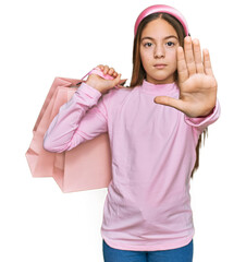 Beautiful brunette little girl holding shopping bags with open hand doing stop sign with serious and confident expression, defense gesture