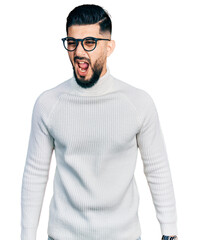Young arab man with beard wearing elegant turtleneck sweater and glasses angry and mad screaming...