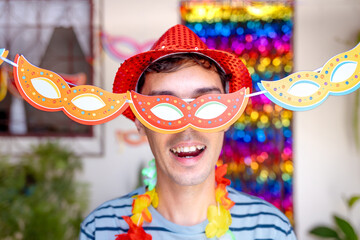 Brazilian man having fun with a carnival mask, set against the backdrop of a house decorated for a...