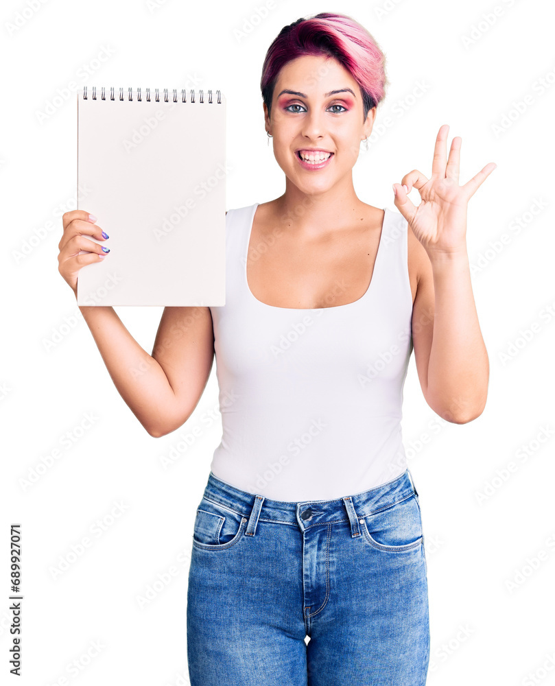 Sticker Young beautiful woman with pink hair holding notebook doing ok sign with fingers, smiling friendly gesturing excellent symbol