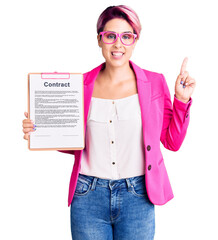 Young beautiful woman with pink hair holding clipboard with contract document with a big smile on face, pointing with hand finger to the side looking at the camera.