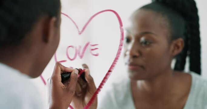 Black woman, mirror and writing heart with lipstick for love, romance or valentines day in bathroom at home. Happy African female person drawing shape, emoji or icon with cosmetics for romantic sign
