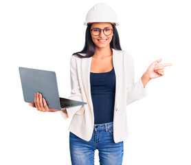 Young beautiful latin girl wearing architect hardhat holding laptop smiling happy pointing with hand and finger to the side
