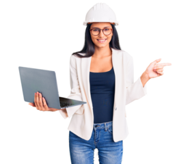 Young beautiful latin girl wearing architect hardhat holding laptop smiling happy pointing with hand and finger to the side