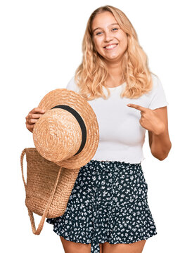 Young blonde girl holding summer hat and wicker handbag pointing finger to one self smiling happy and proud