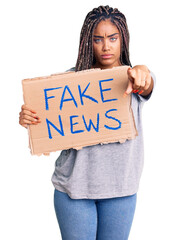 Young african american woman with braids holding fake news banner pointing with finger to the...
