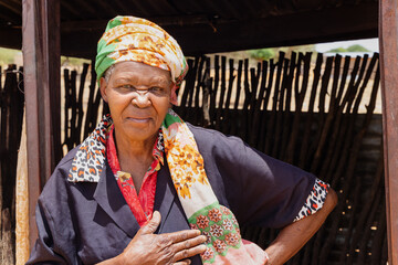 village poor african old woman, wearing a scarf, standing in front of the shed