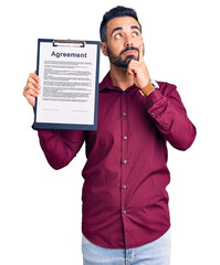 Young hispanic man holding clipboard with agreement document serious face thinking about question...