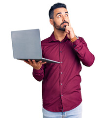 Young hispanic man holding laptop serious face thinking about question with hand on chin,...