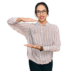 Fototapeta premium Young hispanic girl wearing casual clothes and glasses gesturing with hands showing big and large size sign, measure symbol. smiling looking at the camera. measuring concept.
