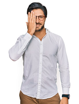 Young hispanic man wearing business shirt and glasses covering one eye with hand, confident smile on face and surprise emotion.