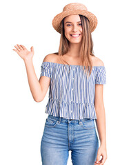 Obraz na płótnie Canvas Young beautiful girl wearing hat and t shirt smiling cheerful presenting and pointing with palm of hand looking at the camera.