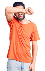 Young handsome man with beard wearing casual t-shirt covering eyes with arm smiling cheerful and funny. blind concept.