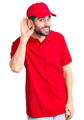 Young handsome man with beard wearing delivery uniform smiling with hand over ear listening an hearing to rumor or gossip. deafness concept.