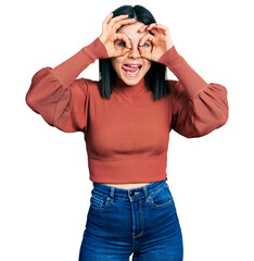 Young brunette woman with blue eyes wearing elegant sweater doing ok gesture like binoculars sticking tongue out, eyes looking through fingers. crazy expression.