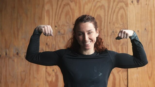 Empowering charm: A smiling strong woman flexes double biceps, radiating strength in a captivating pose. Strong woman smiling at the gym. Beautiful smile. red-head girl doing exercise at CrossFit.