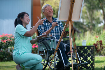 Senior man in wheelchair relaxes his mind by painting on a canvas frame with his caregiver nearby encouraging him, concept of caring for the elderly to be happy