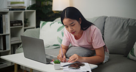Fototapeta na wymiar Portrait of Young Asian woman checks her finances using a calculator on smartphone and makes notes in a notebook while sitting on the sofa at home,Home finance