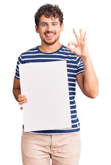 Young handsome man with curly hair holding blank empty banner doing ok sign with fingers, smiling friendly gesturing excellent symbol