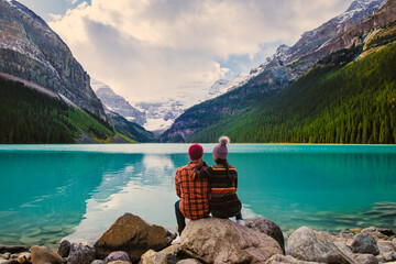 Lake Louise Banff National Park in the Canadian Rocky Mountains. A young couple of men and women sitting on a rock by the lake during a cold day in Autumn in Canada watching the sunset - obrazy, fototapety, plakaty