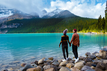Lake Louise Banff National Park in the Canadian Rocky Mountains. A young couple of men and women standing on a rock by the lake during a cold day in Autumn in Canada watching the sunset - obrazy, fototapety, plakaty