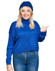 Young caucasian woman wearing wool winter sweater and cap smiling with happy face looking and pointing to the side with thumb up.