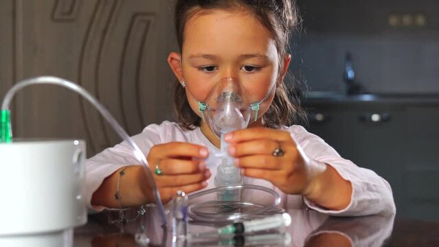 Little child girl undergoing pneumonia treatment at home, breathing through a nebulizer. She sits in a mask, playing with medicines during the process.